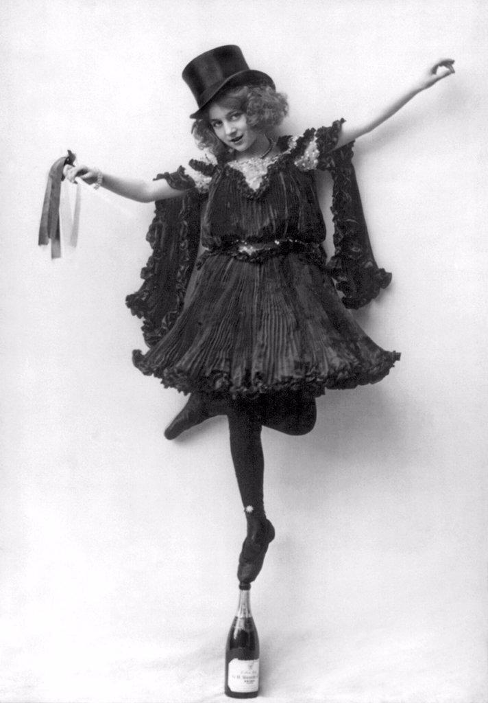A WOMAN IN AN ORNATE COSTUME WITH BLACK PRESSED PLEATS AND A TOP HAT, STANDING EN POINTE ON A CHAMPAGNE BOTTLE