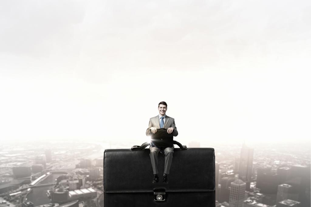 YOUNG SMILING BUSINESSMAN SITTING ON GIANT BRIEFCASE