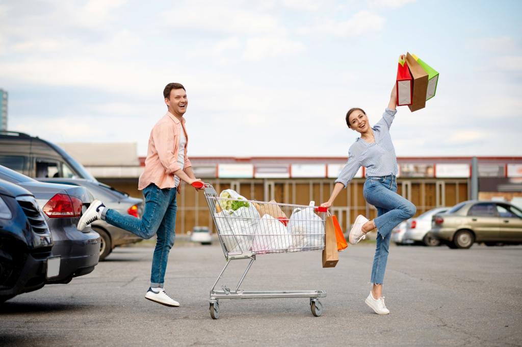 Cheerful family couple with bags in cart on supermarket car parking.