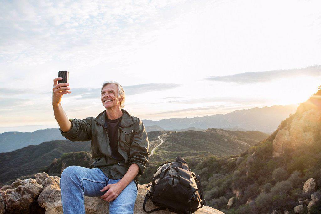 OLDER CAUCASIAN MAN TAKING CELL PHONE PHOTOGRAPH ON ROCKY HILLTOP