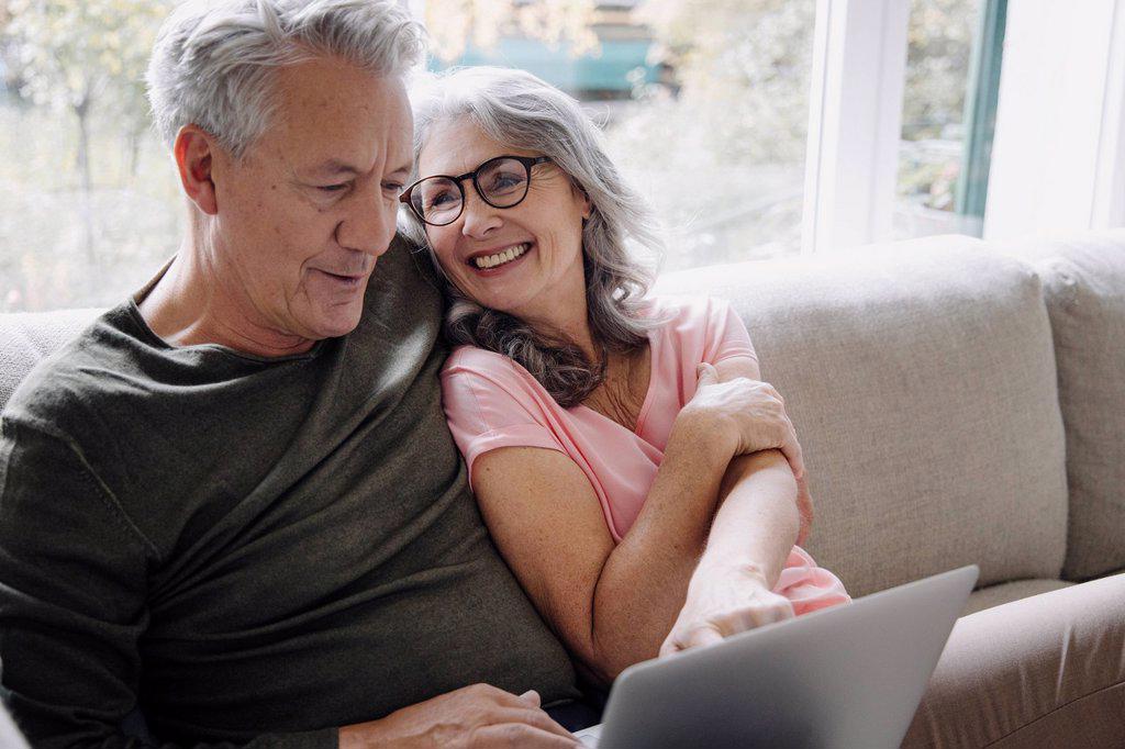 HAPPY SENIOR COUPLE WITH LAPTOP RELAXING ON COUCH AT HOME