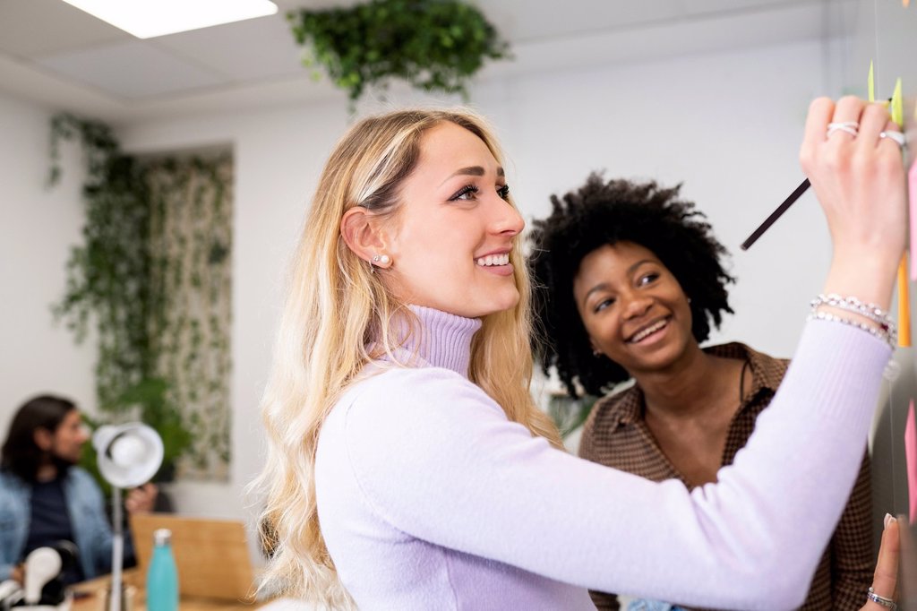 SMILING YOUNG BUSINESSWOMAN WRITING ON ADHESIVE NOTES WHILE DISCUSSING WITH FEMALE COWORKER IN OFFICE