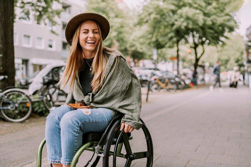 CHEERFUL YOUNG WOMAN SITTING IN WHEELCHAIR AT SIDEWALK