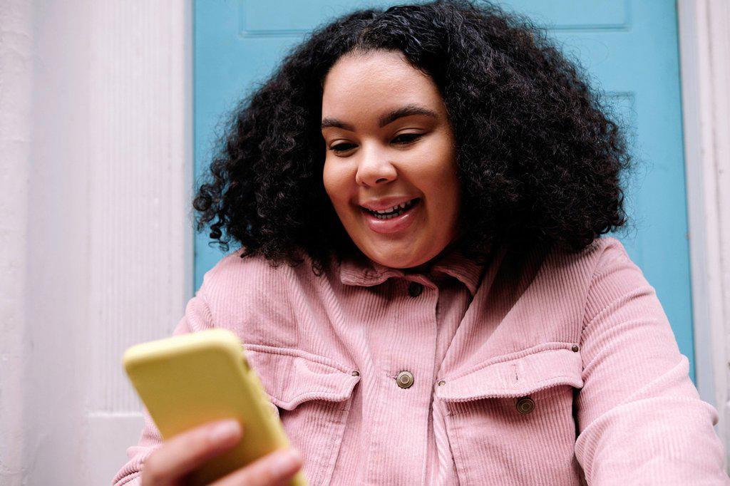 HAPPY YOUNG WOMAN WITH CURLY HAIR SURFING NET THROUGH SMART PHONE