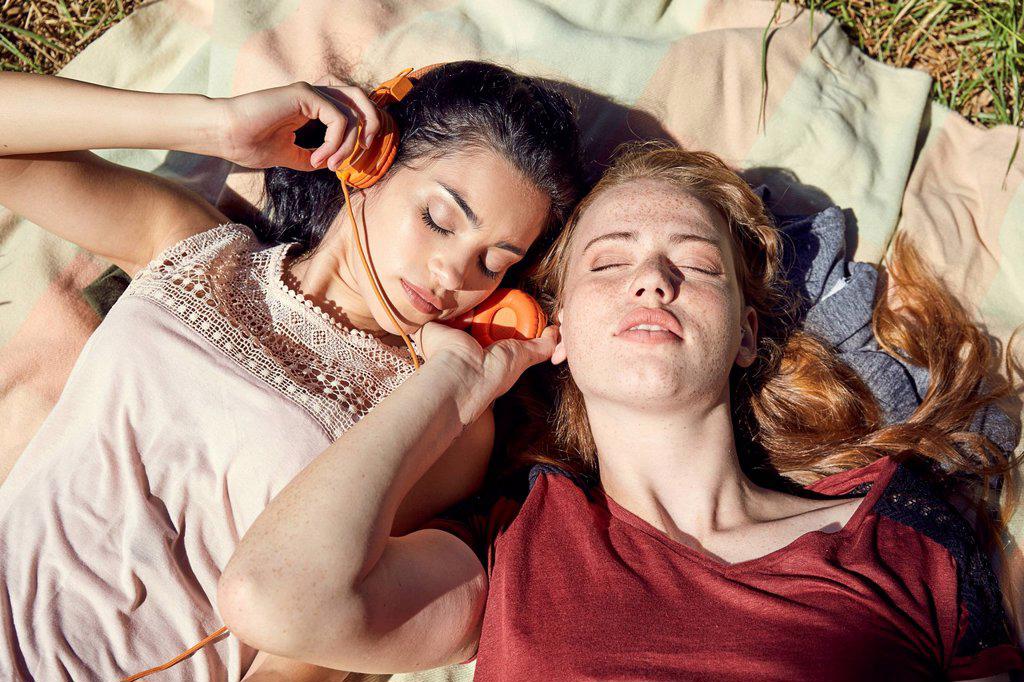 TWO YOUNG WOMEN LYING SIDE BY SIDE ON BLANKET LISTENING MUSIC WITH HEADPHONES