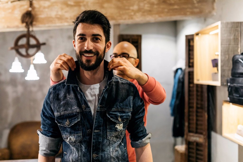 HANDSOME MAN TRYING ON NEW DENIM JACKET WITH STYLIST IN MODERN BOUTIQUE