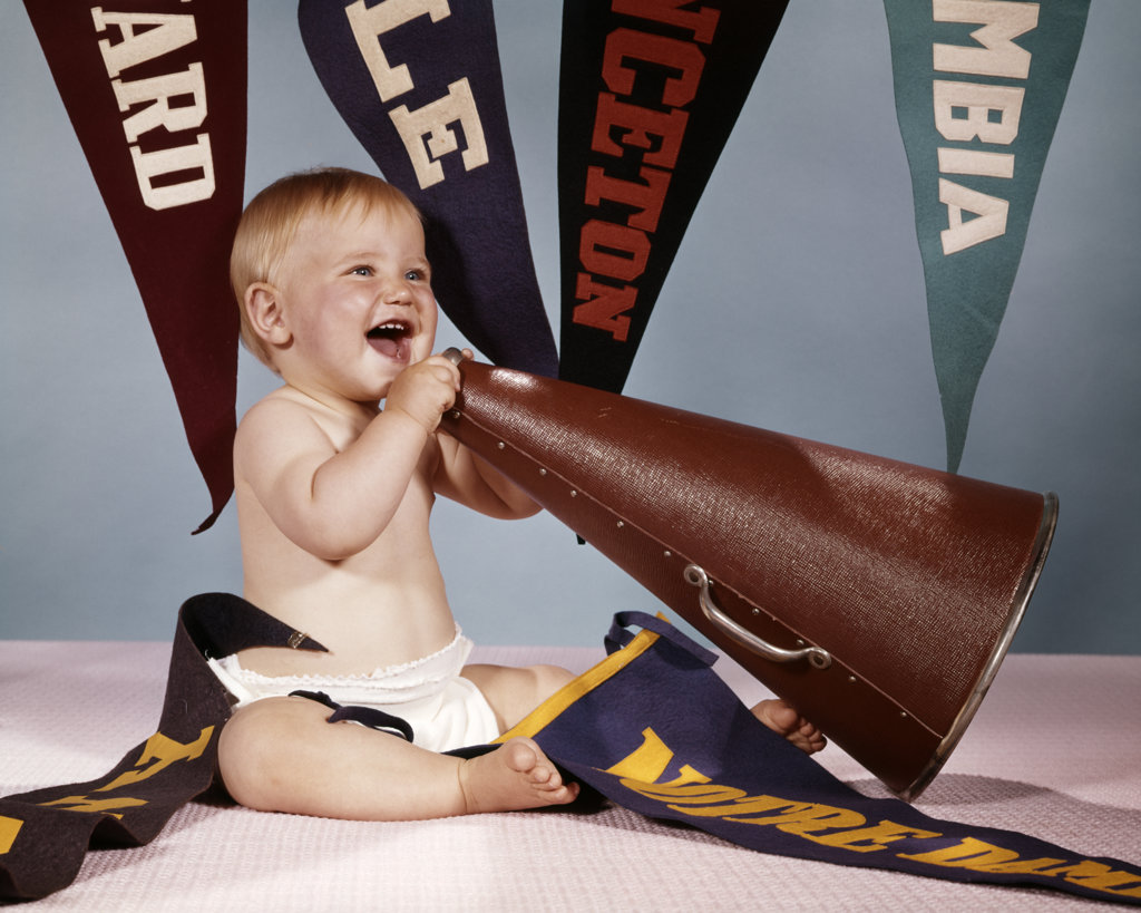 EXCITED BABY GIRL CHEERLEADER SHOUTING INTO MEGAPHONE SURROUNDED BY COLLEGIATE ATHLETIC PENNANTS