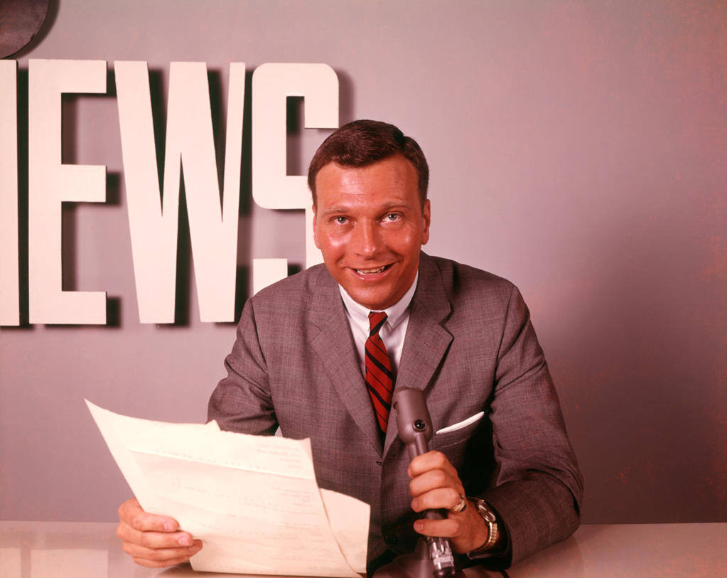 1960S Smiling Broadcast Reporter Newsman Announcer At News Desk Holding Microphone Papers Reading Report