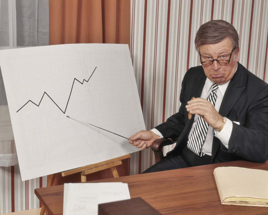UNHAPPY BUSINESSMAN LOOKING AT CAMERA POINTING TO GRAPH CHART INDICATING A DECLINE IN SALES PROFITS BUSINESS