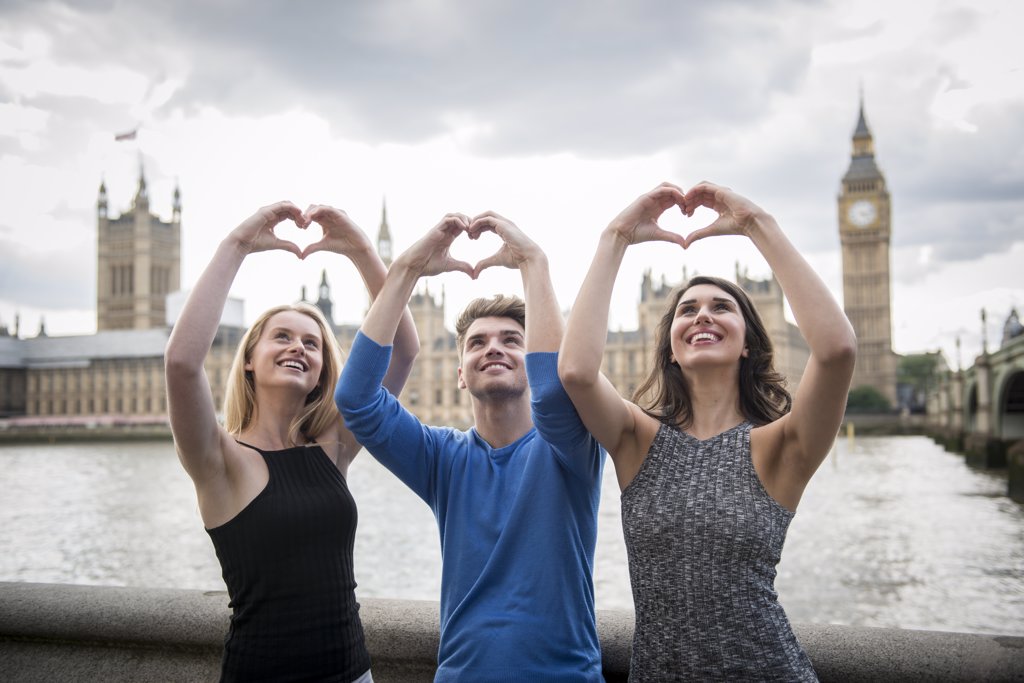 THREE FRIENDS MAKE A HEART SHAPE WITH THEIR HANDS WITH THE HOUSES OF PARLIAMENT IN THE BACKGROUND.