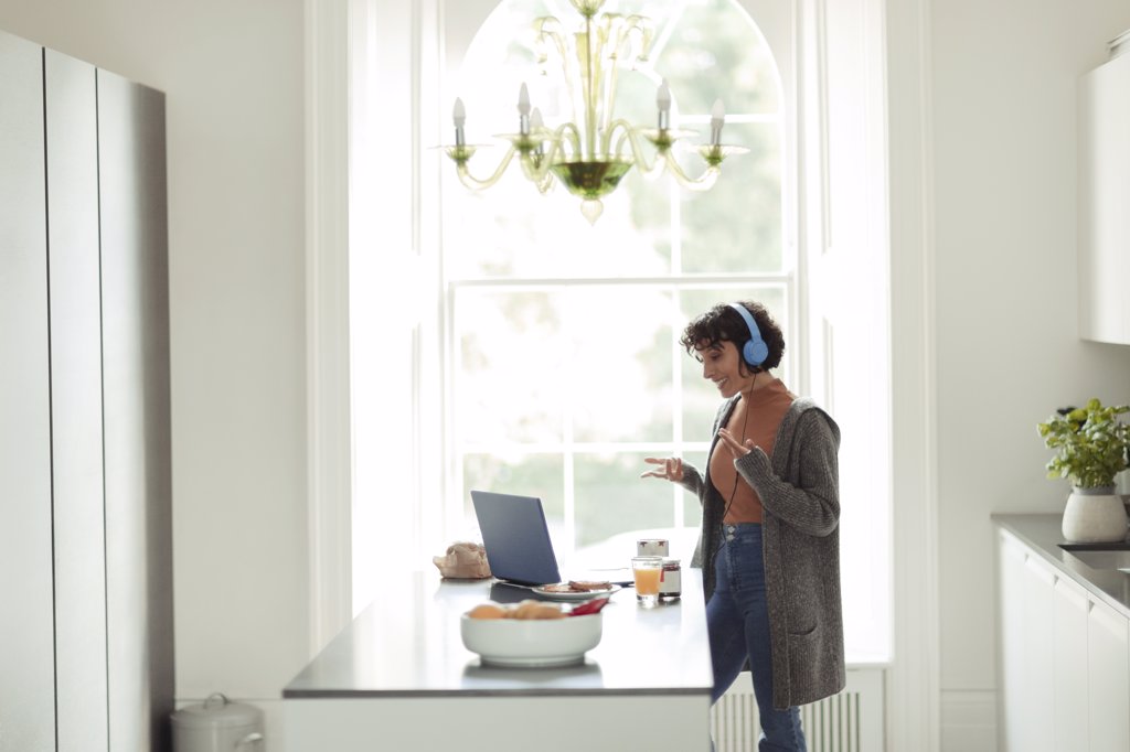 WOMAN WITH HEADPHONES VIDEO CONFERENCING AT LAPTOP IN KITCHEN