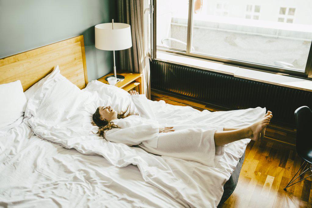 SMILING WOMAN IN BATHROBE HAVING FUN ON BED AT HOTEL ROOM