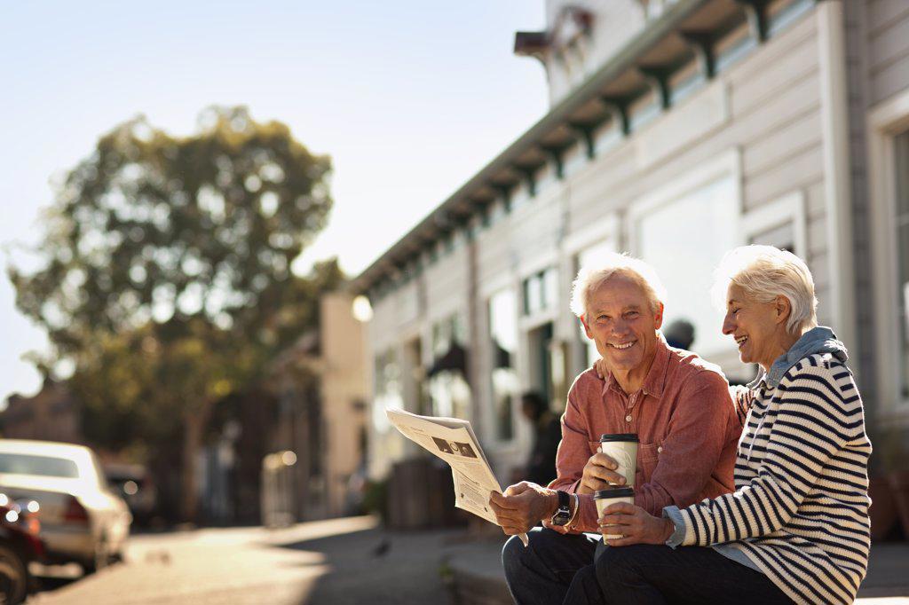 SMILING SENIOR COUPLE SIT READING A NEWSPAPER AND DRINKING COFFEE FROM TAKEAWAY CUPS.