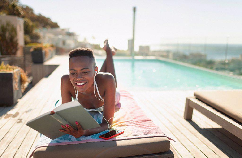 SMILING, CAREFREE YOUNG WOMAN READING BOOK AT SUNNY POOLSIDE