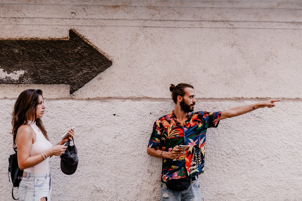 HANDSOME YOUNG BEARDED MAN SHOWING PATH TO THOUGHTFUL BRUNETTE WITH BACKPACK AND SMARTPHONE AGAINST WHITE WALL WITH ARROW