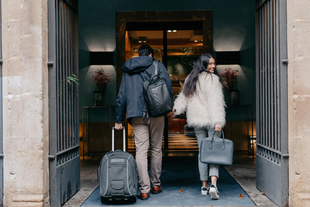 HAPPY STYLISH YOUNG COUPLE IN LOVE WITH SUITCASE AND BAGS HOLDING HANDS AND ENTERING MODERN CITY HOTEL DURING TRAVEL