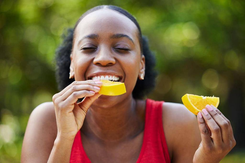 Black woman, vitamin C and eating orange slice for natural nutrition or citrus diet in nature outdoors