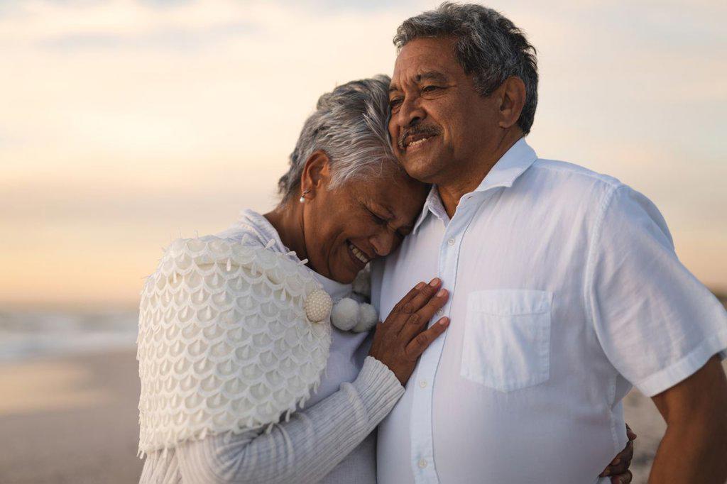 SMILING SENIOR BIRACIAL WOMAN EMBRACING MAN LOOKING AWAY WHILE STANDING AT BEACH DURING SUNSET