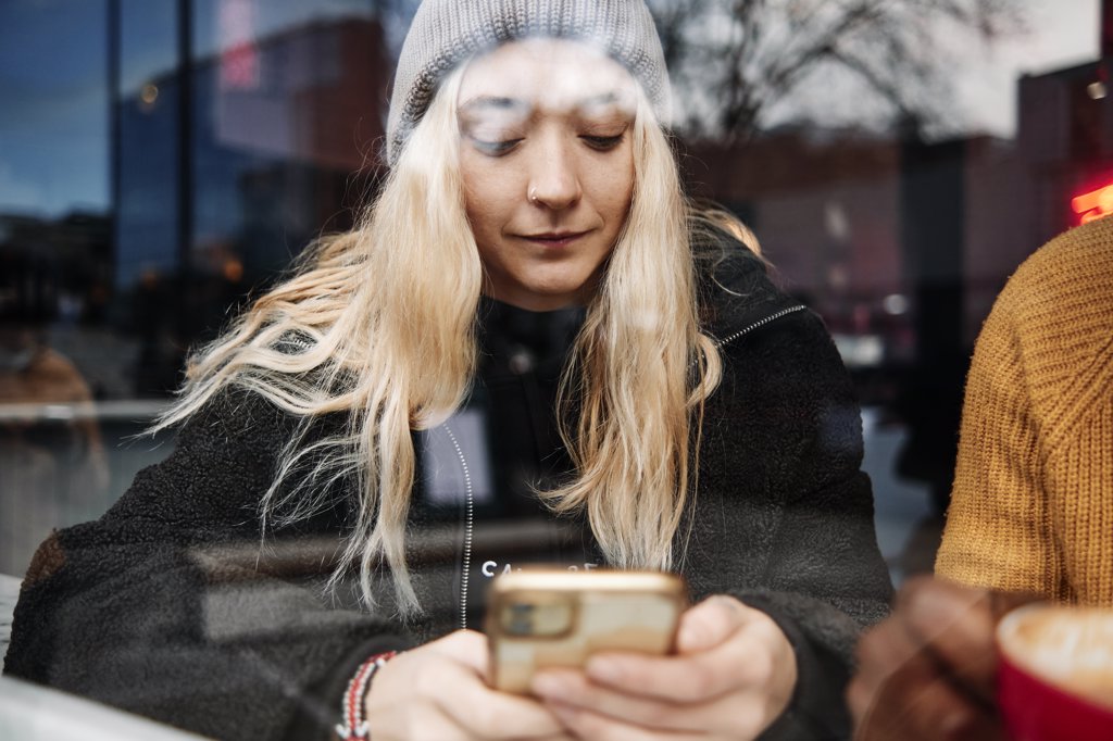 YOUNG FEMALE CHECKING HER PHONE, SITTING AT THE WINDOW OF A CAFE WITH THE CITY REFLECTION ON THE GLASS.