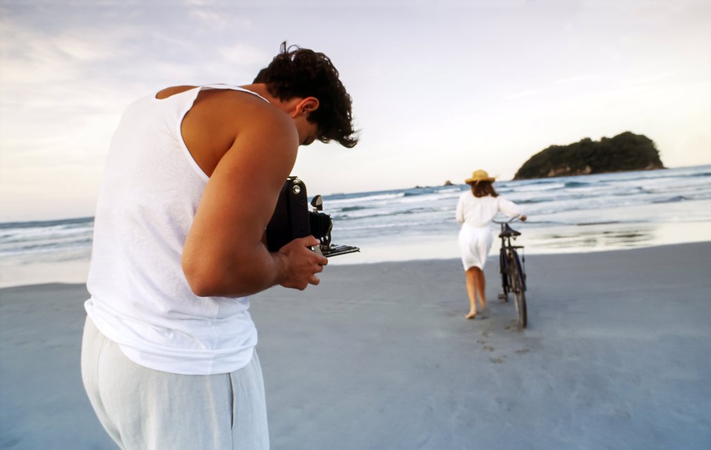 MAN BENT OVER CAMERA TAKING PHOTOS OF WOMAN PUSHING BIKE IN THE SAND AT THE BEACH